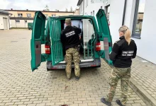 dwóch funkcjonariuszy Straży Granicznej RP (Polish Border Guard) w mundurach polowych. Stoją przy zielonym pojeździe służbowym z otwartymi tylnymi drzwiami, który jest przystosowany do transportu osób zatrzymanych – widoczne są metalowe kraty wewnątrz. Jeden z funkcjonariuszy pomaga osobie wsiąść lub wysiąść z pojazdu. Scena ma miejsce na brukowanym dziedzińcu obok budynku administracyjnego.