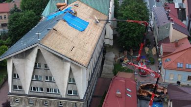 remont dachu dużego budynku, prawdopodobnie kościoła lub budynku użyteczności publicznej. Część starego pokrycia dachowego została już zdjęta, a dach zabezpieczono niebieską plandeką. Prace prowadzone są z użyciem podnośników koszowych, a robotnicy znajdują się zarówno na dachu, jak i w koszach podnośników. Na dole, obok budynku, rozłożone są materiały budowlane, w tym miedziana blacha lub pokrycie dachowe. Otoczenie to centrum małego miasta — widać zabudowę mieszkalną, chodniki i ulicę biegnącą obok budynku.