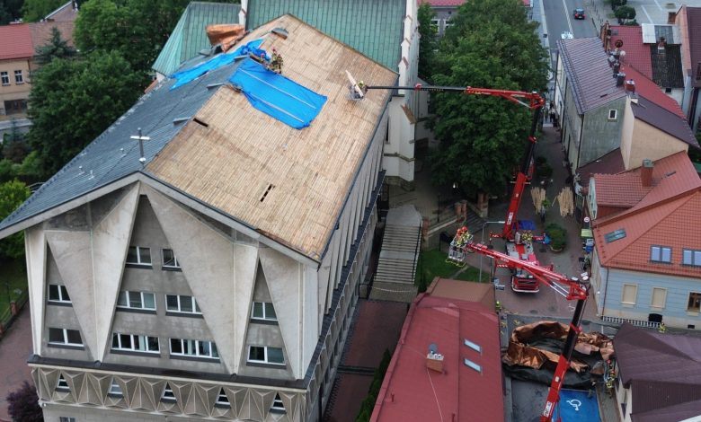 remont dachu dużego budynku, prawdopodobnie kościoła lub budynku użyteczności publicznej. Część starego pokrycia dachowego została już zdjęta, a dach zabezpieczono niebieską plandeką. Prace prowadzone są z użyciem podnośników koszowych, a robotnicy znajdują się zarówno na dachu, jak i w koszach podnośników. Na dole, obok budynku, rozłożone są materiały budowlane, w tym miedziana blacha lub pokrycie dachowe. Otoczenie to centrum małego miasta — widać zabudowę mieszkalną, chodniki i ulicę biegnącą obok budynku.