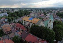 widok z lotu ptaka na centrum małego europejskiego miasta, prawdopodobnie w Polsce. W centrum kadru znajduje się duży budynek kościelny o charakterystycznym dachu, który jest obecnie w trakcie remontu – część pokrycia dachowego została usunięta, a na jednej z połaci widać niebieską plandekę zabezpieczającą. Obok znajduje się biały kościół z zielonym, strzelistym dachem i wieżą z zegarem, co sugeruje, że może to być zabytkowa świątynia. Wokół widzimy gęsto zabudowaną dzielnicę z domami o czerwonych i brązowych dachach. W tle rozciąga się panorama miasta z niską zabudową, licznymi drzewami i ulicami. W oddali widać również przemysłowy komin i dalsze tereny zielone. Zdjęcie zostało wykonane prawdopodobnie po burzy – niebo jest pochmurne i zachmurzone.