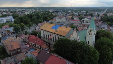 widok z lotu ptaka na centrum małego europejskiego miasta, prawdopodobnie w Polsce. W centrum kadru znajduje się duży budynek kościelny o charakterystycznym dachu, który jest obecnie w trakcie remontu – część pokrycia dachowego została usunięta, a na jednej z połaci widać niebieską plandekę zabezpieczającą. Obok znajduje się biały kościół z zielonym, strzelistym dachem i wieżą z zegarem, co sugeruje, że może to być zabytkowa świątynia. Wokół widzimy gęsto zabudowaną dzielnicę z domami o czerwonych i brązowych dachach. W tle rozciąga się panorama miasta z niską zabudową, licznymi drzewami i ulicami. W oddali widać również przemysłowy komin i dalsze tereny zielone. Zdjęcie zostało wykonane prawdopodobnie po burzy – niebo jest pochmurne i zachmurzone.