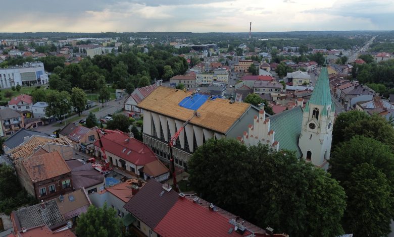 widok z lotu ptaka na centrum małego europejskiego miasta, prawdopodobnie w Polsce. W centrum kadru znajduje się duży budynek kościelny o charakterystycznym dachu, który jest obecnie w trakcie remontu – część pokrycia dachowego została usunięta, a na jednej z połaci widać niebieską plandekę zabezpieczającą. Obok znajduje się biały kościół z zielonym, strzelistym dachem i wieżą z zegarem, co sugeruje, że może to być zabytkowa świątynia. Wokół widzimy gęsto zabudowaną dzielnicę z domami o czerwonych i brązowych dachach. W tle rozciąga się panorama miasta z niską zabudową, licznymi drzewami i ulicami. W oddali widać również przemysłowy komin i dalsze tereny zielone. Zdjęcie zostało wykonane prawdopodobnie po burzy – niebo jest pochmurne i zachmurzone.