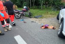 miejsce wypadku drogowego z udziałem motocykla i samochodu osobowego. Na obrazie widoczne są: Motocykl zaparkowany na poboczu drogi. Samochód osobowy z widocznym prawym przednim kołem i otwartymi drzwiami pasażera. Na jezdni i poboczu leżą porozrzucane przedmioty, w tym buty, ubrania, kask i plastikowe elementy – prawdopodobnie pochodzące z pojazdów lub należące do uczestników wypadku. Na ziemi leży osoba okryta kocem termicznym, co sugeruje, że mogła zostać poszkodowana. Obok stoją ratownicy medyczni ubrani w charakterystyczne czerwono-czarne stroje. Tło pokazuje zalesiony teren oraz zaparkowane rowery i budynek. Sytuacja wygląda na poważną, a służby ratunkowe są już na miejscu.
