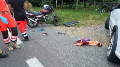 miejsce wypadku drogowego z udziałem motocykla i samochodu osobowego. Na obrazie widoczne są: Motocykl zaparkowany na poboczu drogi. Samochód osobowy z widocznym prawym przednim kołem i otwartymi drzwiami pasażera. Na jezdni i poboczu leżą porozrzucane przedmioty, w tym buty, ubrania, kask i plastikowe elementy – prawdopodobnie pochodzące z pojazdów lub należące do uczestników wypadku. Na ziemi leży osoba okryta kocem termicznym, co sugeruje, że mogła zostać poszkodowana. Obok stoją ratownicy medyczni ubrani w charakterystyczne czerwono-czarne stroje. Tło pokazuje zalesiony teren oraz zaparkowane rowery i budynek. Sytuacja wygląda na poważną, a służby ratunkowe są już na miejscu.