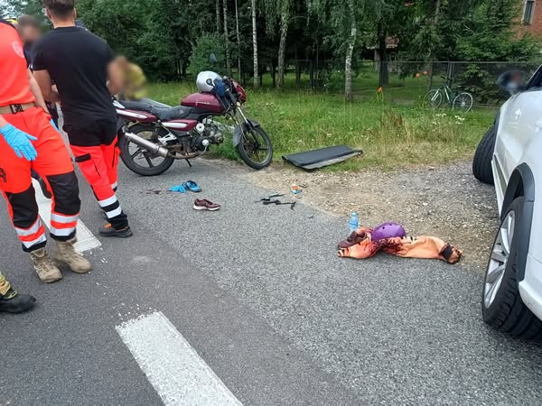 miejsce wypadku drogowego z udziałem motocykla i samochodu osobowego. Na obrazie widoczne są: Motocykl zaparkowany na poboczu drogi. Samochód osobowy z widocznym prawym przednim kołem i otwartymi drzwiami pasażera. Na jezdni i poboczu leżą porozrzucane przedmioty, w tym buty, ubrania, kask i plastikowe elementy – prawdopodobnie pochodzące z pojazdów lub należące do uczestników wypadku. Na ziemi leży osoba okryta kocem termicznym, co sugeruje, że mogła zostać poszkodowana. Obok stoją ratownicy medyczni ubrani w charakterystyczne czerwono-czarne stroje. Tło pokazuje zalesiony teren oraz zaparkowane rowery i budynek. Sytuacja wygląda na poważną, a służby ratunkowe są już na miejscu.