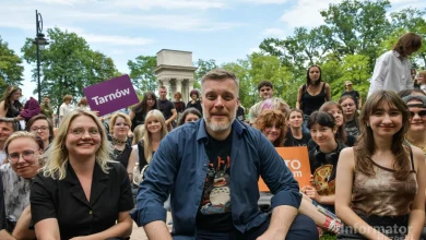 widzimy grupę uśmiechniętych ludzi zgromadzonych na świeżym powietrzu, prawdopodobnie podczas wydarzenia społecznego lub kulturalnego. W centrum siedzi mężczyzna z siwiejącą brodą i koszulką z postacią Totoro z japońskiego filmu animowanego. Wokół niego siedzą młode osoby, niektóre trzymają tabliczki z nazwami miast, np. „Tarnów” i „TO my”. W tle widoczna jest kolumnada oraz zieleń drzew, sugerująca parkową scenerię. Atmosfera jest swobodna i przyjazna, a całość ma charakter integracyjny lub aktywistyczny. W prawym dolnym rogu znajduje się logo „Informator Brzeski”.