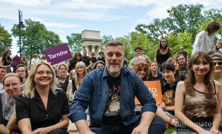 widzimy grupę uśmiechniętych ludzi zgromadzonych na świeżym powietrzu, prawdopodobnie podczas wydarzenia społecznego lub kulturalnego. W centrum siedzi mężczyzna z siwiejącą brodą i koszulką z postacią Totoro z japońskiego filmu animowanego. Wokół niego siedzą młode osoby, niektóre trzymają tabliczki z nazwami miast, np. „Tarnów” i „TO my”. W tle widoczna jest kolumnada oraz zieleń drzew, sugerująca parkową scenerię. Atmosfera jest swobodna i przyjazna, a całość ma charakter integracyjny lub aktywistyczny. W prawym dolnym rogu znajduje się logo „Informator Brzeski”.