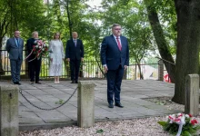 Grupa sześciu osób w formalnych strojach podczas uroczystości upamiętniającej na świeżym powietrzu. Jeden mężczyzna stoi z przodu, reszta – w tym kobieta w białej sukience – stoi z tyłu. Przed nimi, na ziemi, leży wieniec z biało-czerwoną wstążką. W tle drzewa i metalowe ogrodzenie.
