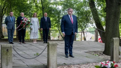 Grupa sześciu osób w formalnych strojach podczas uroczystości upamiętniającej na świeżym powietrzu. Jeden mężczyzna stoi z przodu, reszta – w tym kobieta w białej sukience – stoi z tyłu. Przed nimi, na ziemi, leży wieniec z biało-czerwoną wstążką. W tle drzewa i metalowe ogrodzenie.