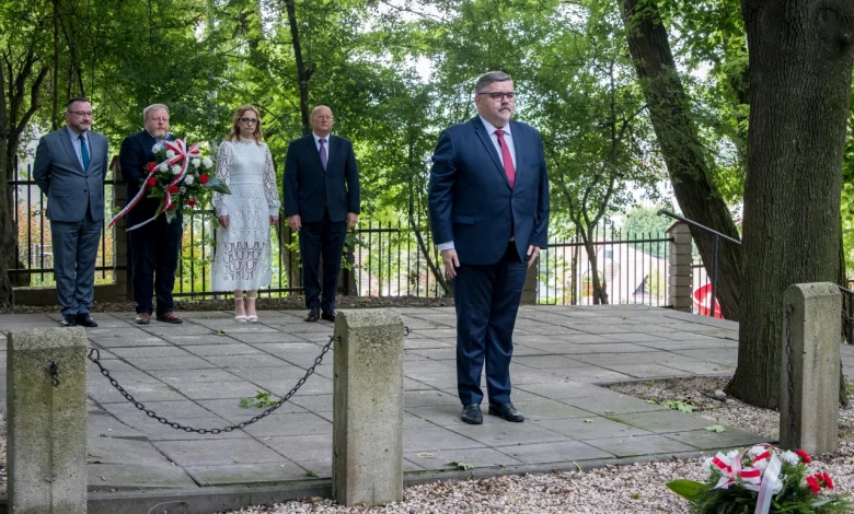 Grupa sześciu osób w formalnych strojach podczas uroczystości upamiętniającej na świeżym powietrzu. Jeden mężczyzna stoi z przodu, reszta – w tym kobieta w białej sukience – stoi z tyłu. Przed nimi, na ziemi, leży wieniec z biało-czerwoną wstążką. W tle drzewa i metalowe ogrodzenie.