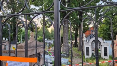 Zdjęcie przedstawia cmentarz widziany przez zamkniętą bramę z kutego żelaza. Wewnątrz widoczne są groby, płyty nagrobne oraz liczne drzewa. Na bramie zawieszono pomarańczową taśmę ostrzegawczą oraz kartkę z informacją w języku polskim. Cmentarz został prawdopodobnie tymczasowo zamknięty — być może z powodu niebezpieczeństwa związanego z drzewami (np. ryzyko przewrócenia się). Na kartce znajduje się fragment ostrzeżenia: „PO OGLĘDZINACH KOMUNALNYCH ... STWIERDZA PRZESŁANKĘ, KTÓRA JEDNOZNACZNIE WSKAZUJE NA KONIECZNOŚĆ ZABEZPIECZENIA ... DRZEW ... ZAGROŻENIA ŻYCIA LUDZKIEGO.” Oznacza to, że na podstawie oględzin stwierdzono zagrożenie związane z drzewami, które może stanowić niebezpieczeństwo dla ludzi. W związku z tym miejsce zostało zabezpieczone i prawdopodobnie wyłączone z użytkowania do czasu usunięcia zagrożenia.