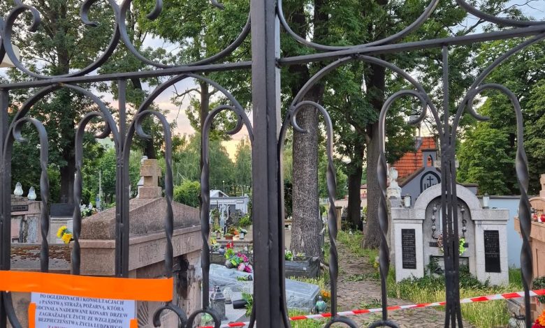 Zdjęcie przedstawia cmentarz widziany przez zamkniętą bramę z kutego żelaza. Wewnątrz widoczne są groby, płyty nagrobne oraz liczne drzewa. Na bramie zawieszono pomarańczową taśmę ostrzegawczą oraz kartkę z informacją w języku polskim. Cmentarz został prawdopodobnie tymczasowo zamknięty — być może z powodu niebezpieczeństwa związanego z drzewami (np. ryzyko przewrócenia się). Na kartce znajduje się fragment ostrzeżenia: „PO OGLĘDZINACH KOMUNALNYCH ... STWIERDZA PRZESŁANKĘ, KTÓRA JEDNOZNACZNIE WSKAZUJE NA KONIECZNOŚĆ ZABEZPIECZENIA ... DRZEW ... ZAGROŻENIA ŻYCIA LUDZKIEGO.” Oznacza to, że na podstawie oględzin stwierdzono zagrożenie związane z drzewami, które może stanowić niebezpieczeństwo dla ludzi. W związku z tym miejsce zostało zabezpieczone i prawdopodobnie wyłączone z użytkowania do czasu usunięcia zagrożenia.
