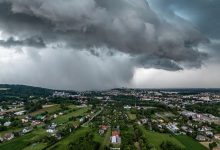 burzowa chmura w kształcie leja z intensywnym opadem deszczu, zbliżająca się do miasta. Widok z lotu ptaka ukazuje zabudowania mieszkalne i zielone tereny, kontrastujące z ciemnym, groźnym niebem. Chmura burzowa dominuje nad krajobrazem, zwiastując nadchodzącą nawałnicę.