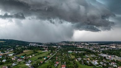 burzowa chmura w kształcie leja z intensywnym opadem deszczu, zbliżająca się do miasta. Widok z lotu ptaka ukazuje zabudowania mieszkalne i zielone tereny, kontrastujące z ciemnym, groźnym niebem. Chmura burzowa dominuje nad krajobrazem, zwiastując nadchodzącą nawałnicę.