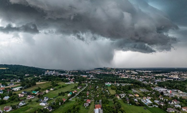 burzowa chmura w kształcie leja z intensywnym opadem deszczu, zbliżająca się do miasta. Widok z lotu ptaka ukazuje zabudowania mieszkalne i zielone tereny, kontrastujące z ciemnym, groźnym niebem. Chmura burzowa dominuje nad krajobrazem, zwiastując nadchodzącą nawałnicę.