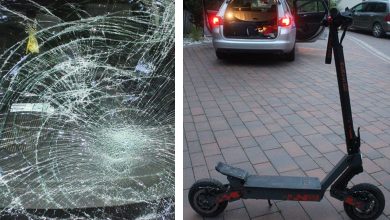 Po lewej stronie zdjęcia widać pękniętą i mocno rozbitą szybę samochodu – pajęczyna pęknięć wskazuje na silne uderzenie. Po prawej stronie zdjęcia znajduje się hulajnoga elektryczna ustawiona na kostce brukowej, w tle otwarty bagażnik srebrnego samochodu, prawdopodobnie związanego ze zdarzeniem.