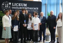 grupa uczniów i dorosłych stojących wspólnie przed tablicą z napisem „Liceum Ogólnokształcące im. Mikołaja Kopernika w Brzesku”. Kilkoro uczniów trzyma dyplomy oraz puchar, co sugeruje, że zostali wyróżnieni lub nagrodzeni w jakimś konkursie lub wydarzeniu szkolnym. Wszyscy są elegancko ubrani, co świadczy o uroczystym charakterze spotkania. Tło stanowi czarna tablica z logo szkoły oraz niebieskie rolety i rośliny doniczkowe.