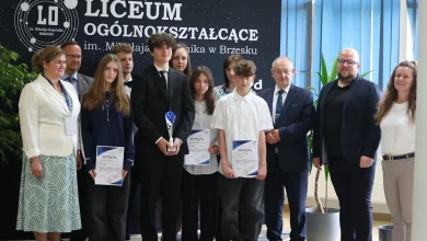 grupa uczniów i dorosłych stojących wspólnie przed tablicą z napisem „Liceum Ogólnokształcące im. Mikołaja Kopernika w Brzesku”. Kilkoro uczniów trzyma dyplomy oraz puchar, co sugeruje, że zostali wyróżnieni lub nagrodzeni w jakimś konkursie lub wydarzeniu szkolnym. Wszyscy są elegancko ubrani, co świadczy o uroczystym charakterze spotkania. Tło stanowi czarna tablica z logo szkoły oraz niebieskie rolety i rośliny doniczkowe.