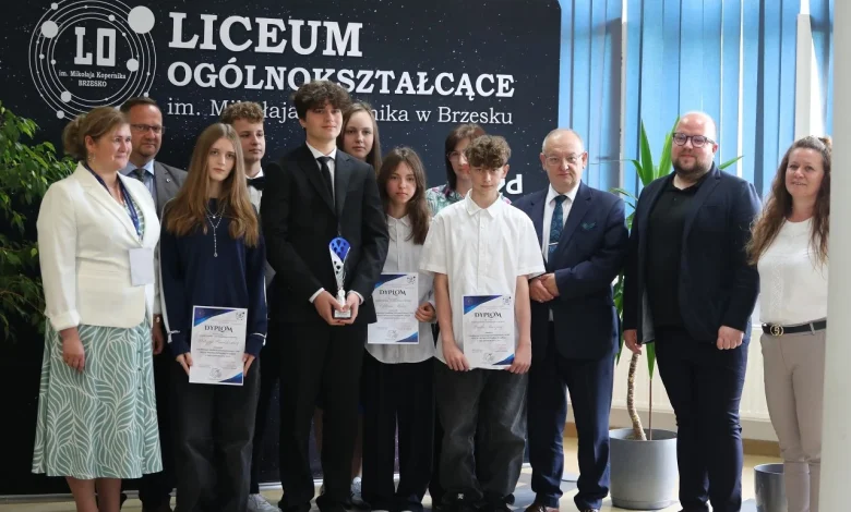 grupa uczniów i dorosłych stojących wspólnie przed tablicą z napisem „Liceum Ogólnokształcące im. Mikołaja Kopernika w Brzesku”. Kilkoro uczniów trzyma dyplomy oraz puchar, co sugeruje, że zostali wyróżnieni lub nagrodzeni w jakimś konkursie lub wydarzeniu szkolnym. Wszyscy są elegancko ubrani, co świadczy o uroczystym charakterze spotkania. Tło stanowi czarna tablica z logo szkoły oraz niebieskie rolety i rośliny doniczkowe.