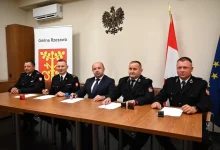 Mężczyźni siedzą za stołem, przed sobą kartki papieru, za nim herb gminy, godło i flagi