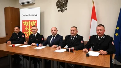 Mężczyźni siedzą za stołem, przed sobą kartki papieru, za nim herb gminy, godło i flagi