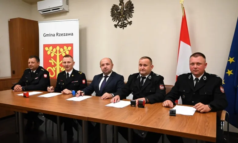 Mężczyźni siedzą za stołem, przed sobą kartki papieru, za nim herb gminy, godło i flagi