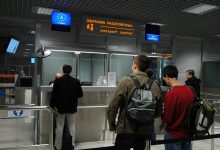 punkt kontroli paszportowej na lotnisku. Kilka osób stoi w kolejce, czekając na odprawę paszportową. Nad stanowiskami widoczne są znaki w języku polskim i angielskim: „ODPRAWA PASZPORTOWA / passport control”. Jeden z napisów wskazuje stanowisko dla obywateli Unii Europejskiej (oznaczone symbolem UE), a drugie dla wszystkich paszportów („ALL PASSPORTS”). W tle znajdują się funkcjonariusze straży granicznej oraz elementy infrastruktury lotniskowej.