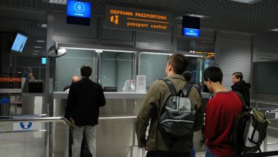 punkt kontroli paszportowej na lotnisku. Kilka osób stoi w kolejce, czekając na odprawę paszportową. Nad stanowiskami widoczne są znaki w języku polskim i angielskim: „ODPRAWA PASZPORTOWA / passport control”. Jeden z napisów wskazuje stanowisko dla obywateli Unii Europejskiej (oznaczone symbolem UE), a drugie dla wszystkich paszportów („ALL PASSPORTS”). W tle znajdują się funkcjonariusze straży granicznej oraz elementy infrastruktury lotniskowej.