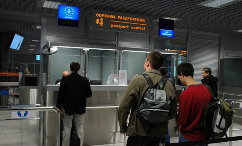 punkt kontroli paszportowej na lotnisku. Kilka osób stoi w kolejce, czekając na odprawę paszportową. Nad stanowiskami widoczne są znaki w języku polskim i angielskim: „ODPRAWA PASZPORTOWA / passport control”. Jeden z napisów wskazuje stanowisko dla obywateli Unii Europejskiej (oznaczone symbolem UE), a drugie dla wszystkich paszportów („ALL PASSPORTS”). W tle znajdują się funkcjonariusze straży granicznej oraz elementy infrastruktury lotniskowej.