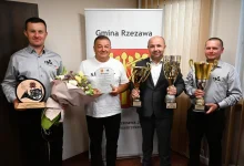czterech mężczyzn stojących w pomieszczeniu, pozujących do zdjęcia z trofeami i dyplomami. W tle znajduje się baner z napisem „Gmina Rzezawa”, co wskazuje, że wydarzenie miało miejsce w tej gminie, położonej w województwie małopolskim. Mężczyzna po lewej trzyma statuetkę oraz bukiet kwiatów. Drugi od lewej, w białej koszulce, trzyma dyplom i również bukiet kwiatów. Trzeci, ubrany w garnitur, trzyma dwa duże puchary. Ostatni mężczyzna również dzierży okazały puchar. Wygląda na to, że są to członkowie zespołu, który odniósł sukces — prawdopodobnie strażacy ochotnicy (na koszulach widnieje herb OSP – Ochotniczej Straży Pożarnej). Nagrody i puchary mogą być efektem zwycięstwa w zawodach strażackich, zawodach sportowych lub innego prestiżowego konkursu. Atmosfera zdjęcia jest radosna i oficjalna.