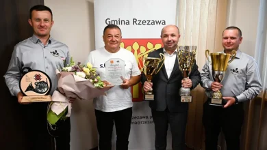 czterech mężczyzn stojących w pomieszczeniu, pozujących do zdjęcia z trofeami i dyplomami. W tle znajduje się baner z napisem „Gmina Rzezawa”, co wskazuje, że wydarzenie miało miejsce w tej gminie, położonej w województwie małopolskim. Mężczyzna po lewej trzyma statuetkę oraz bukiet kwiatów. Drugi od lewej, w białej koszulce, trzyma dyplom i również bukiet kwiatów. Trzeci, ubrany w garnitur, trzyma dwa duże puchary. Ostatni mężczyzna również dzierży okazały puchar. Wygląda na to, że są to członkowie zespołu, który odniósł sukces — prawdopodobnie strażacy ochotnicy (na koszulach widnieje herb OSP – Ochotniczej Straży Pożarnej). Nagrody i puchary mogą być efektem zwycięstwa w zawodach strażackich, zawodach sportowych lub innego prestiżowego konkursu. Atmosfera zdjęcia jest radosna i oficjalna.