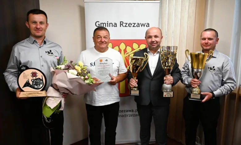 czterech mężczyzn stojących w pomieszczeniu, pozujących do zdjęcia z trofeami i dyplomami. W tle znajduje się baner z napisem „Gmina Rzezawa”, co wskazuje, że wydarzenie miało miejsce w tej gminie, położonej w województwie małopolskim. Mężczyzna po lewej trzyma statuetkę oraz bukiet kwiatów. Drugi od lewej, w białej koszulce, trzyma dyplom i również bukiet kwiatów. Trzeci, ubrany w garnitur, trzyma dwa duże puchary. Ostatni mężczyzna również dzierży okazały puchar. Wygląda na to, że są to członkowie zespołu, który odniósł sukces — prawdopodobnie strażacy ochotnicy (na koszulach widnieje herb OSP – Ochotniczej Straży Pożarnej). Nagrody i puchary mogą być efektem zwycięstwa w zawodach strażackich, zawodach sportowych lub innego prestiżowego konkursu. Atmosfera zdjęcia jest radosna i oficjalna.