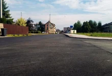 Nowo wyasfaltowana droga w dzielnicy mieszkalnej. W tle widać domy, zieleń i częściowo zachmurzone niebo.
