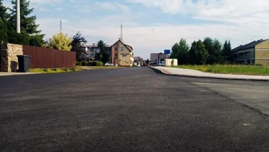 Nowo wyasfaltowana droga w dzielnicy mieszkalnej. W tle widać domy, zieleń i częściowo zachmurzone niebo.