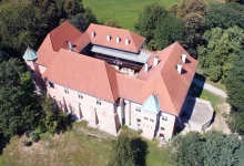 Zamek z czerwonym dachem, otoczony zielenią i drzewami. Budowla ma kształt zbliżony do prostokąta z wewnętrznym dziedzińcem, co sugeruje, że jest to zamek typu czworobocznego. Dachy są strome, kryte czerwoną dachówką, a naroża budowli są zaakcentowane wieżyczkami z niebiesko-zielonymi hełmami. Zamek wykonany jest z kamienia lub cegły, a elewacje pokazują elementy typowe dla architektury średniowiecznej lub renesansowej. Budowla znajduje się w lesistej okolicy z otwartą przestrzenią w tle.