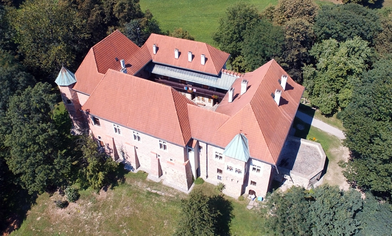 Zamek z czerwonym dachem, otoczony zielenią i drzewami. Budowla ma kształt zbliżony do prostokąta z wewnętrznym dziedzińcem, co sugeruje, że jest to zamek typu czworobocznego. Dachy są strome, kryte czerwoną dachówką, a naroża budowli są zaakcentowane wieżyczkami z niebiesko-zielonymi hełmami. Zamek wykonany jest z kamienia lub cegły, a elewacje pokazują elementy typowe dla architektury średniowiecznej lub renesansowej. Budowla znajduje się w lesistej okolicy z otwartą przestrzenią w tle.