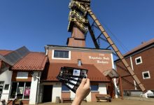 wejście do Kopalni Soli Bochnia, jednej z najstarszych kopalni soli w Europie, założonej w 1248 roku. Budynek ma czerwony dach i jasne ściany, a na elewacji widnieje napis: "Kopalnia Soli Bochnia 1248". Na pierwszym planie ktoś trzyma bilety w dłoni, co sugeruje, że zaraz rozpocznie się zwiedzanie tego zabytkowego obiektu. W tle widać szyb kopalniany z charakterystyczną, wysoką konstrukcją stalową. Nad kopalnią niebo jest bezchmurne i niebieskie, co wskazuje na słoneczny dzień. To miejsce znajduje się w Bochni, w województwie małopolskim, i jest wpisane na Listę Światowego Dziedzictwa UNESCO. Kopalnia oferuje atrakcje takie jak podziemne trasy turystyczne, kaplice wykute w soli, zjazdy windą oraz noclegi w podziemnych komorach.