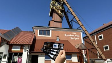 wejście do Kopalni Soli Bochnia, jednej z najstarszych kopalni soli w Europie, założonej w 1248 roku. Budynek ma czerwony dach i jasne ściany, a na elewacji widnieje napis: "Kopalnia Soli Bochnia 1248". Na pierwszym planie ktoś trzyma bilety w dłoni, co sugeruje, że zaraz rozpocznie się zwiedzanie tego zabytkowego obiektu. W tle widać szyb kopalniany z charakterystyczną, wysoką konstrukcją stalową. Nad kopalnią niebo jest bezchmurne i niebieskie, co wskazuje na słoneczny dzień. To miejsce znajduje się w Bochni, w województwie małopolskim, i jest wpisane na Listę Światowego Dziedzictwa UNESCO. Kopalnia oferuje atrakcje takie jak podziemne trasy turystyczne, kaplice wykute w soli, zjazdy windą oraz noclegi w podziemnych komorach.