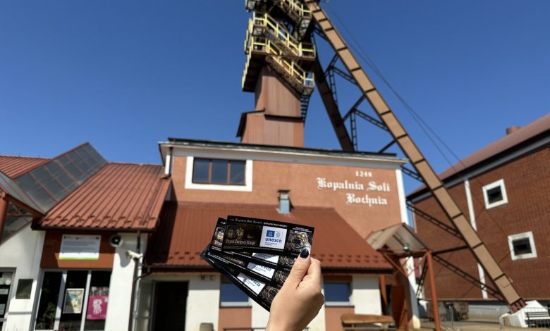 wejście do Kopalni Soli Bochnia, jednej z najstarszych kopalni soli w Europie, założonej w 1248 roku. Budynek ma czerwony dach i jasne ściany, a na elewacji widnieje napis: "Kopalnia Soli Bochnia 1248". Na pierwszym planie ktoś trzyma bilety w dłoni, co sugeruje, że zaraz rozpocznie się zwiedzanie tego zabytkowego obiektu. W tle widać szyb kopalniany z charakterystyczną, wysoką konstrukcją stalową. Nad kopalnią niebo jest bezchmurne i niebieskie, co wskazuje na słoneczny dzień. To miejsce znajduje się w Bochni, w województwie małopolskim, i jest wpisane na Listę Światowego Dziedzictwa UNESCO. Kopalnia oferuje atrakcje takie jak podziemne trasy turystyczne, kaplice wykute w soli, zjazdy windą oraz noclegi w podziemnych komorach.
