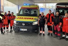 Grupa ratowników medycznych i pracowników pogotowia pozuje do zdjęcia przed dwoma karetkami pogotowia. Osoby ubrane są w czerwono-czarne i pomarańczowe stroje ratownicze. W centrum znajduje się nowoczesna żółto-czerwona karetka Volkswagen z widocznym napisem „AMBULANS” na masce oraz tablicą rejestracyjną „KBC 0533A”. Po prawej stronie stoi druga karetka w klasycznych biało-czerwonych barwach. Zdjęcie wykonano pod zadaszeniem, w tle widoczne są budynki i ogrodzenie.