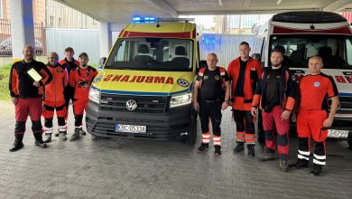 Grupa ratowników medycznych i pracowników pogotowia pozuje do zdjęcia przed dwoma karetkami pogotowia. Osoby ubrane są w czerwono-czarne i pomarańczowe stroje ratownicze. W centrum znajduje się nowoczesna żółto-czerwona karetka Volkswagen z widocznym napisem „AMBULANS” na masce oraz tablicą rejestracyjną „KBC 0533A”. Po prawej stronie stoi druga karetka w klasycznych biało-czerwonych barwach. Zdjęcie wykonano pod zadaszeniem, w tle widoczne są budynki i ogrodzenie.