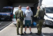 Dwóch umundurowanych funkcjonariuszy prowadzi mężczyznę w szarej bluzie i zielonych spodenkach w stronę białego samochodu dostawczego. W tle widoczna jest karetka pogotowia i inne pojazdy na parkingu.