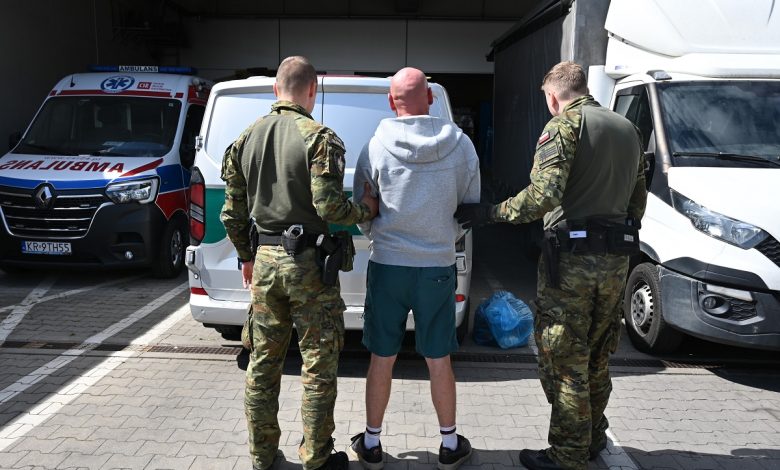 Dwóch umundurowanych funkcjonariuszy prowadzi mężczyznę w szarej bluzie i zielonych spodenkach w stronę białego samochodu dostawczego. W tle widoczna jest karetka pogotowia i inne pojazdy na parkingu.