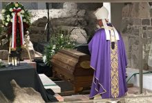 Biskup w fioletowej szacie liturgicznej stoi przed drewnianą trumną ozdobioną kwiatami podczas ceremonii pogrzebowej w kamiennym wnętrzu. Obok trumny znajduje się wieniec z biało-czerwonymi wstążkami.