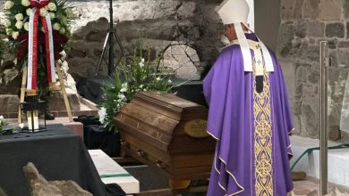 Biskup w fioletowej szacie liturgicznej stoi przed drewnianą trumną ozdobioną kwiatami podczas ceremonii pogrzebowej w kamiennym wnętrzu. Obok trumny znajduje się wieniec z biało-czerwonymi wstążkami.