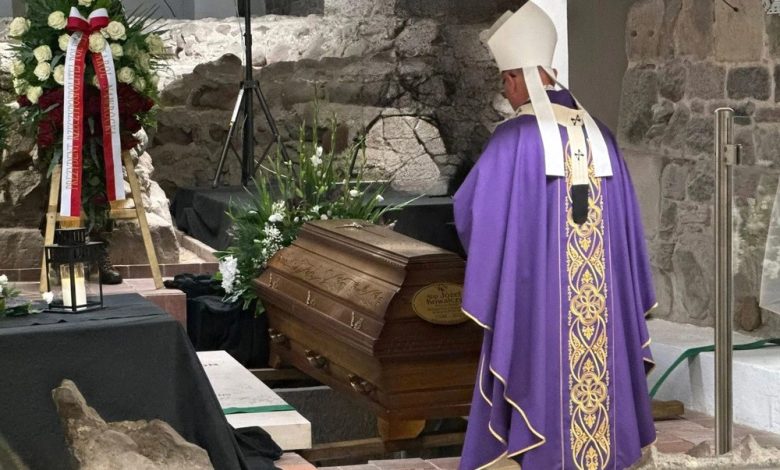 Biskup w fioletowej szacie liturgicznej stoi przed drewnianą trumną ozdobioną kwiatami podczas ceremonii pogrzebowej w kamiennym wnętrzu. Obok trumny znajduje się wieniec z biało-czerwonymi wstążkami.