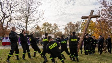Grupa strażaków w czarnych mundurach z napisem „STRAŻ” wspólnie podnosi duży drewniany krzyż za pomocą lin na trawiastym placu. W tle stoją uczestnicy uroczystości, w tym osoby w strojach liturgicznych. Scena odbywa się w jesiennej scenerii, wśród drzew z kolorowymi liśćmi.