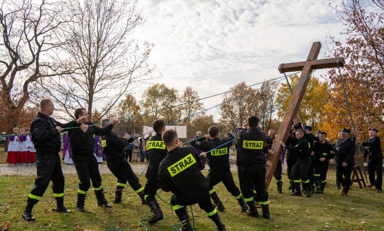 Grupa strażaków w czarnych mundurach z napisem „STRAŻ” wspólnie podnosi duży drewniany krzyż za pomocą lin na trawiastym placu. W tle stoją uczestnicy uroczystości, w tym osoby w strojach liturgicznych. Scena odbywa się w jesiennej scenerii, wśród drzew z kolorowymi liśćmi.