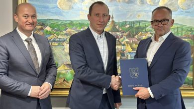 Na zdjęciu widać trzech mężczyzn ubranych w eleganckie garnitury, stojących na tle dużego, kolorowego obrazu przedstawiającego malownicze miasteczko. Dwaj mężczyźni po lewej stronie podają sobie dłonie w geście uścisku, uśmiechając się. Trzeci mężczyzna, stojący po prawej, trzyma w rękach granatową teczkę z herbem i patrzy w stronę aparatu. Wszystkie osoby pozują w formalnej, oficjalnej atmosferze.