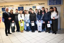 Na zdjęciu widać grupę młodzieży – uczniów wraz z opiekunami – pozujących wewnątrz budynku Urzędu Gminy Dębno. Osoby stoją w jednym rzędzie, część z nich trzyma papierowe torby z logo gminy. Po bokach grupy znajdują się dorośli – kobieta i mężczyzna w formalnych strojach. W tle widoczne są tablice informacyjne oraz plansze z informacjami o środkach finansowych pozyskanych przez urząd. Scena ma oficjalny, ale przyjazny charakter i dokumentuje wizytę młodzieży w urzędzie.